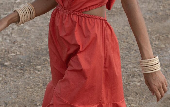 model walking on the beach in a red Johanna Ortiz cut out midi dress with bone colored bangles stacked on both arms and a thin gold chain